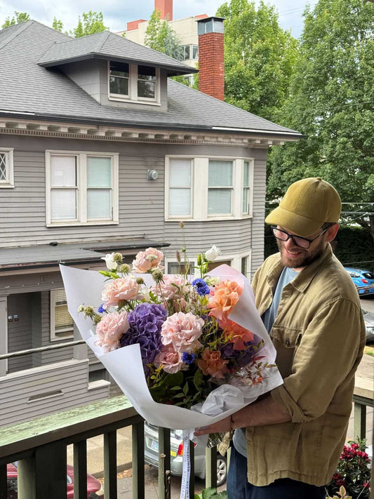 Happy customer holding a large fresh seasonal bouquet with garden roses, purple hydrangea and cornflowers on a Portland porch, by Kvetka Flower Portland