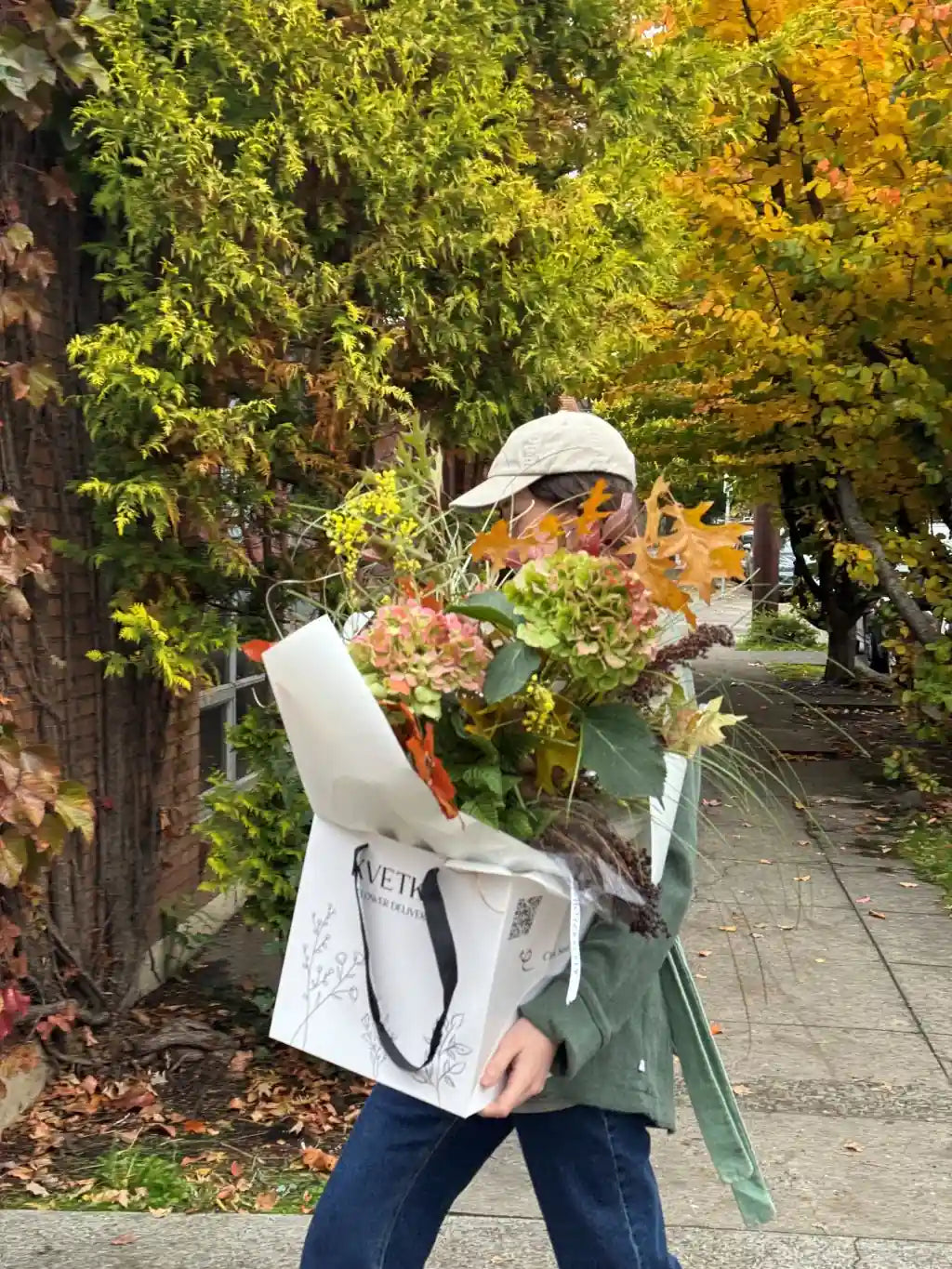 Florist from Kvetka Flower holding a large bouquet of flowers and a white bag with text in an outdoor setting.