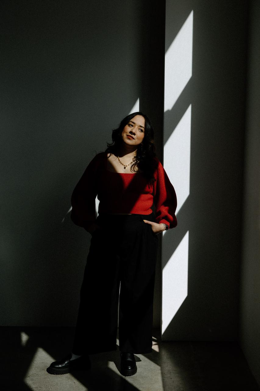 Woman in a red sweater and black pants standing against a wall with light and shadow patterns.