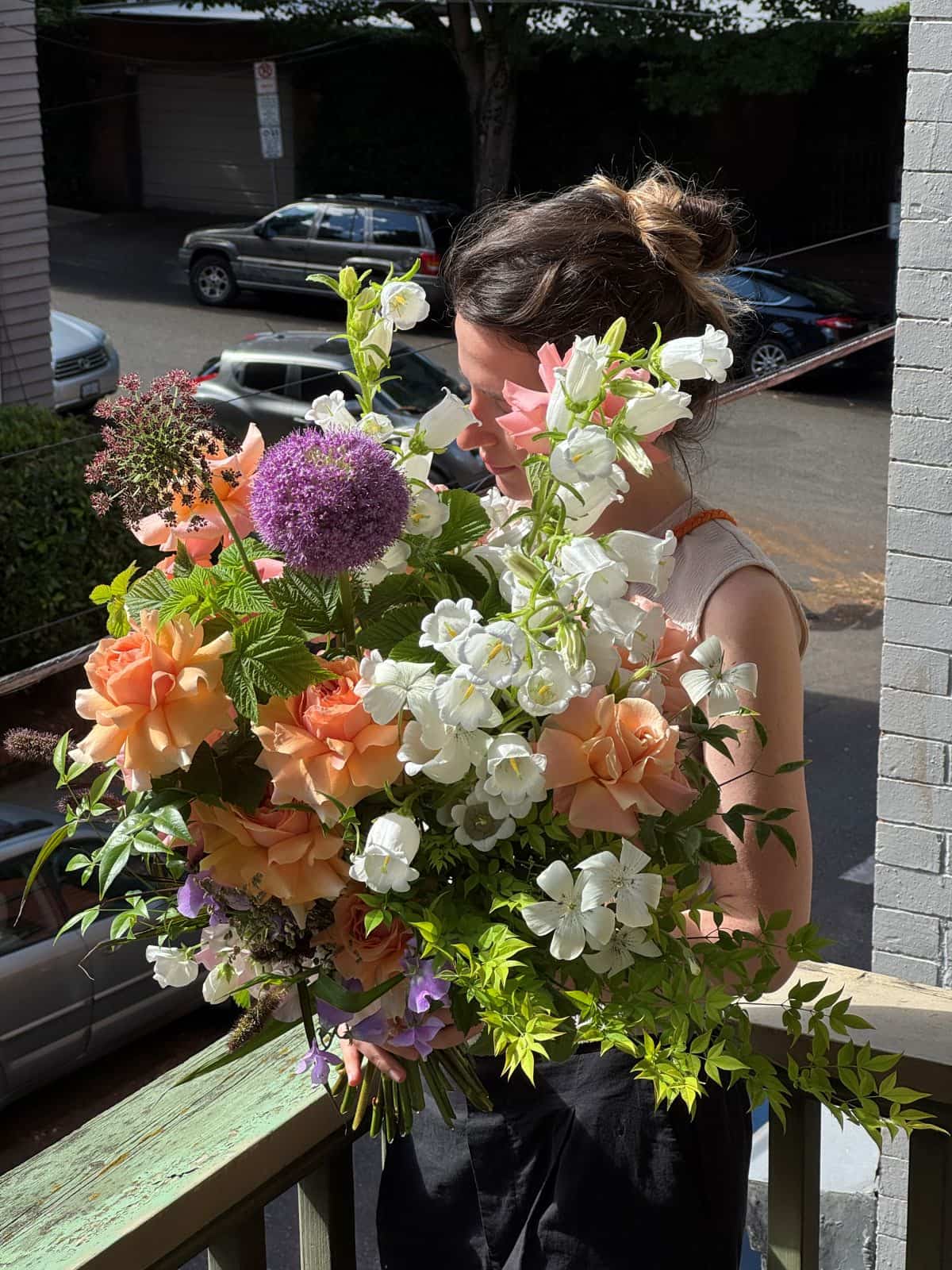 Florist holding an extra large hand-tied bouquet with peach garden roses, white campanula bells and purple allium on a sunny Portland porch, by Kvetka Flower Portland