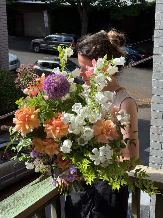 Florist holding an extra large hand-tied bouquet with peach garden roses, white campanula bells and purple allium on a sunny Portland porch, by Kvetka Flower Portland