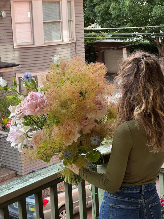 Florist holding a grande hand-tied bouquet with airy smoke bush, pink garden roses, lilies and blue cornflowers on a Portland porch, by Kvetka Flower Portland