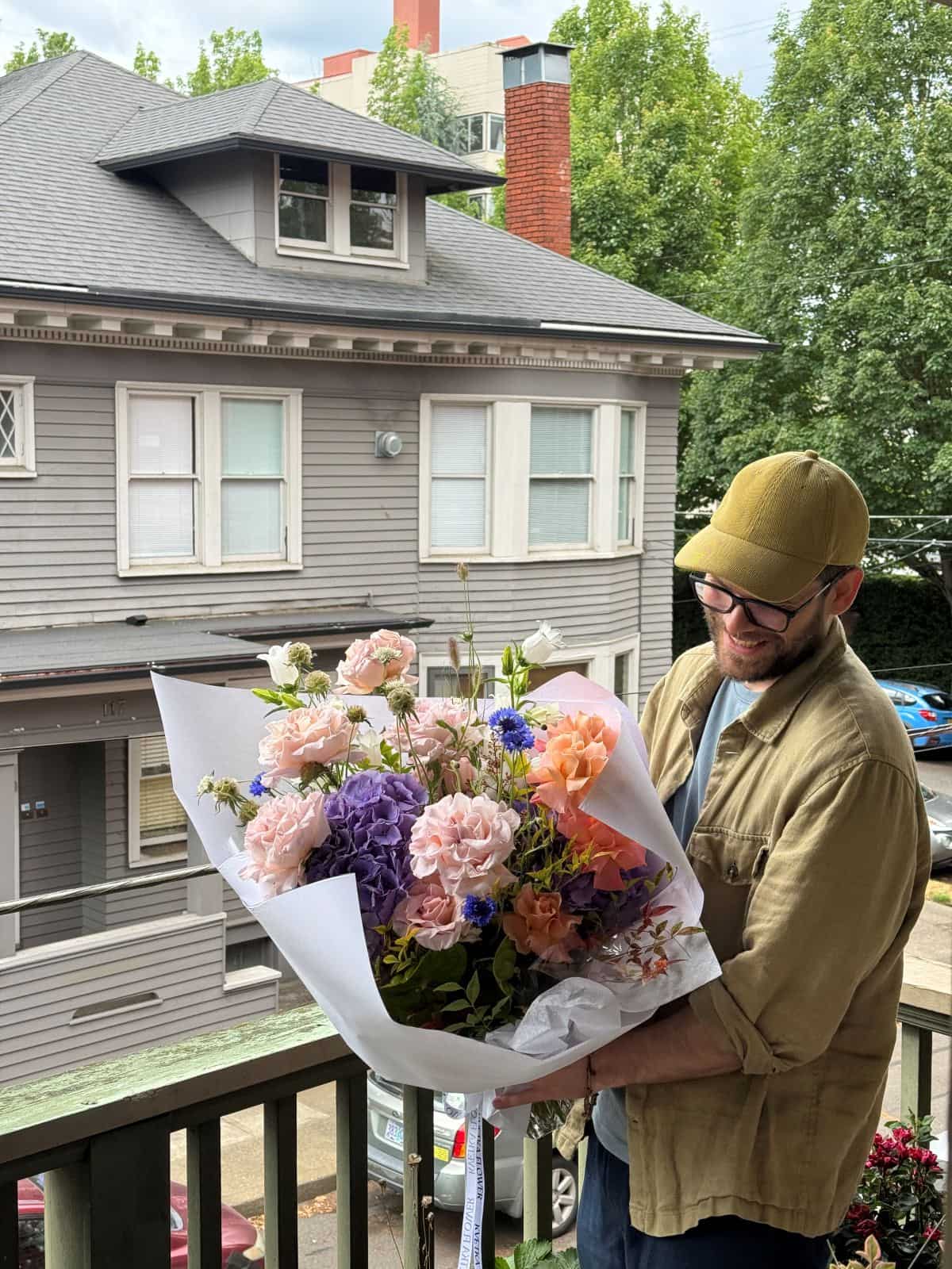 Happy customer holding a large fresh seasonal bouquet with garden roses, purple hydrangea and cornflowers on a Portland porch, by Kvetka Flower Portland
