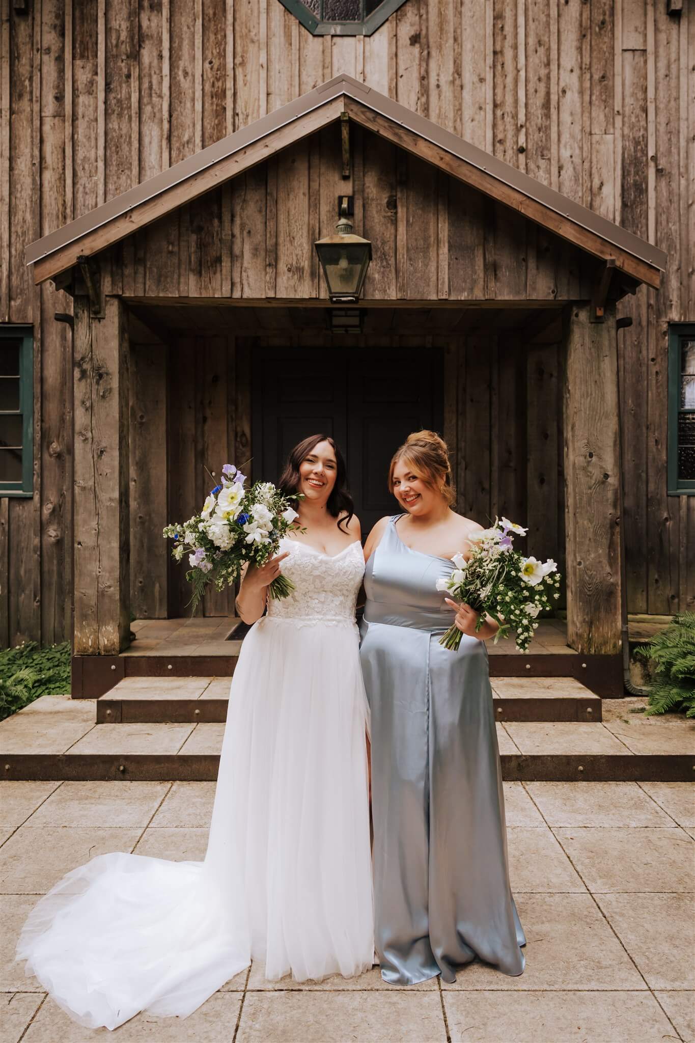 Bride and bridesmaid holding bouquets outside rustic chapel at Camp Colton – by Kvetka Flower, Portland OR
