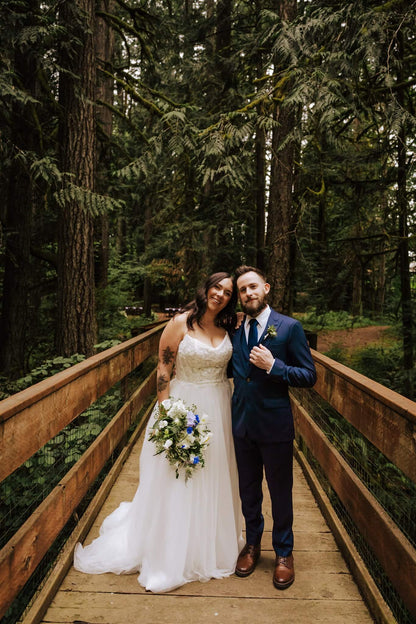 Bride and groom on wooden bridge surrounded by tall fir trees at Camp Colton – by Kvetka Flower, Portland OR

