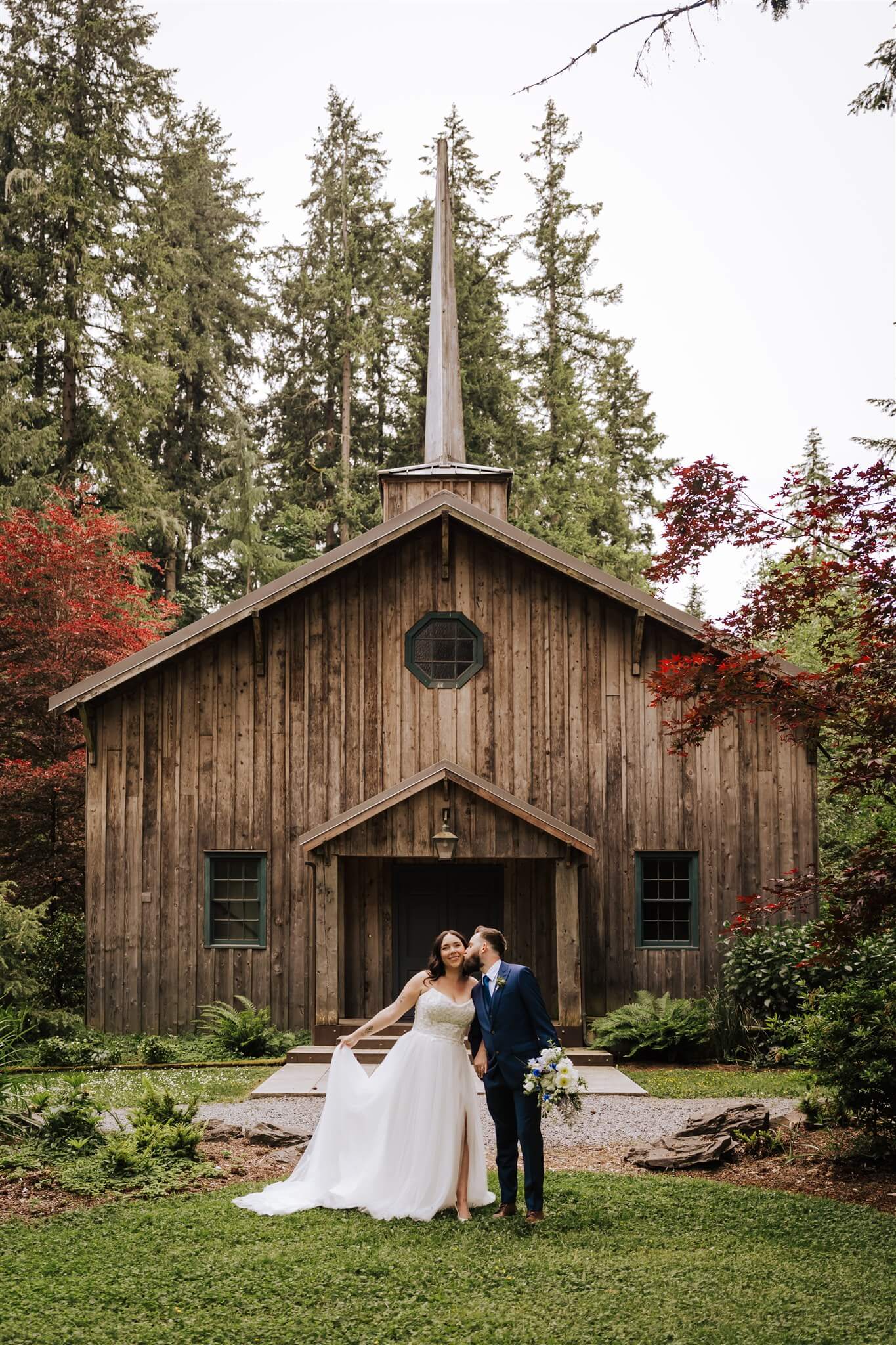Bride and groom sharing a kiss in front of rustic wooden chapel at Camp Colton – by Kvetka Flower, Portland OR

