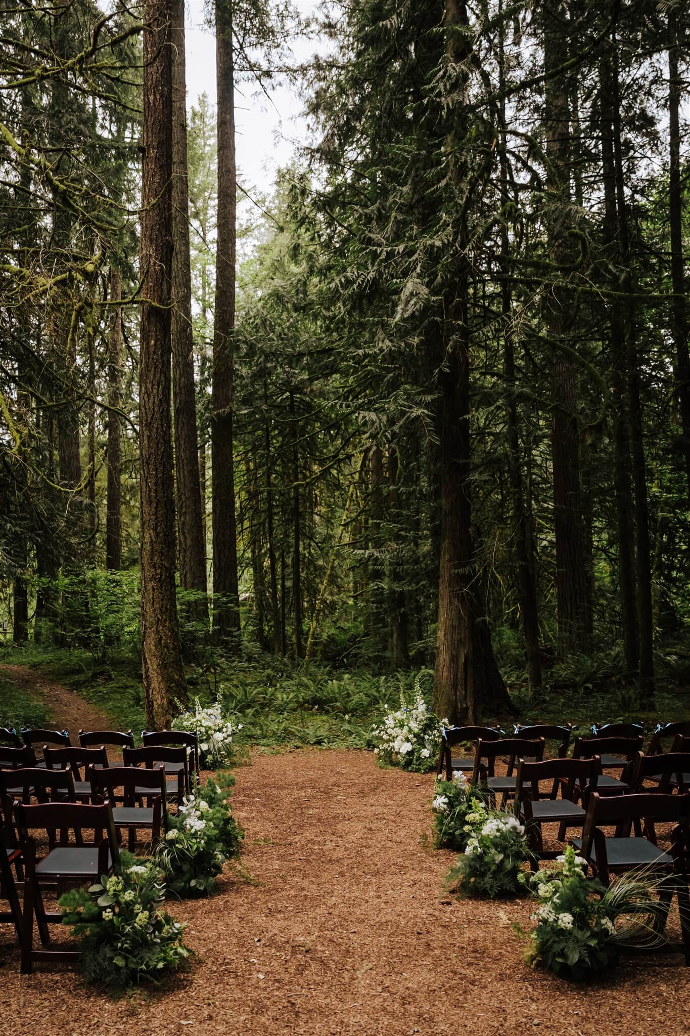 Forest ceremony setup with white flowers and greenery lining the aisle at Camp Colton – by Kvetka Flower, Portland OR