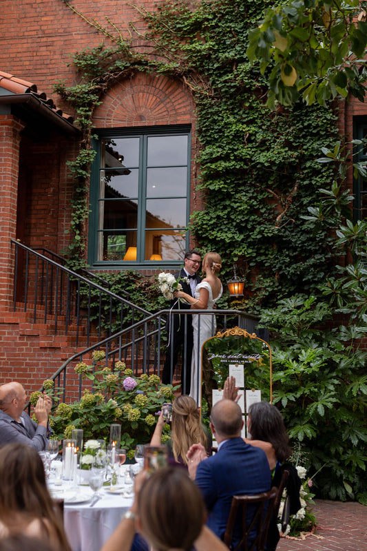 Wedding ceremony taking place outdoors with a couple standing on a balcony, surrounded by guests and greenery.