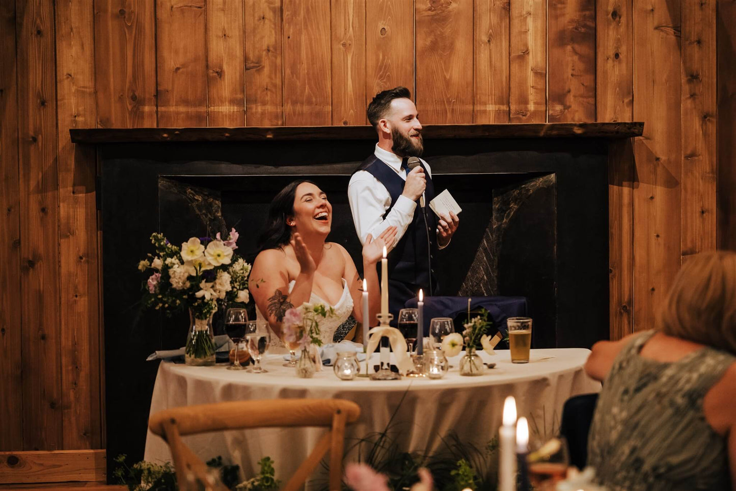 Bride laughing while groom gives toast at sweetheart table with floral decor – by Kvetka Flower, Portland OR
